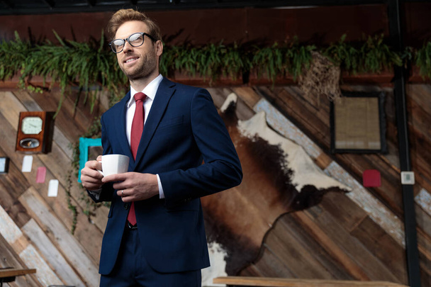 νεαρός επιχειρηματίας με κοστούμι κοιτάζει αλλού και πίνει καφέ, στέκεται σε ένα καφενείο - Φωτογραφία, εικόνα