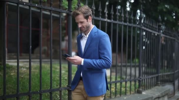 young businessman wearing navy suit walking with hand in pocket while texting on phone and then looking around outside next to a fence - Filmati, video