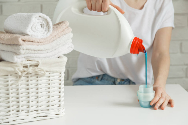Close up of female hands pouring liquid laundry detergent into cap on white rustic table with towels on background in bathroom - Foto, Imagen