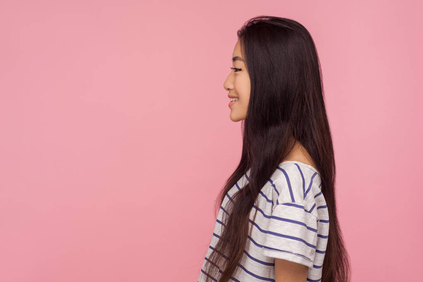 Side view of cheerful pretty asian girl with long brunette hair in striped t-shirt looking at copy space with smile, enjoying happy life, good mood. indoor studio shot isolated on pink background - Foto, Imagen