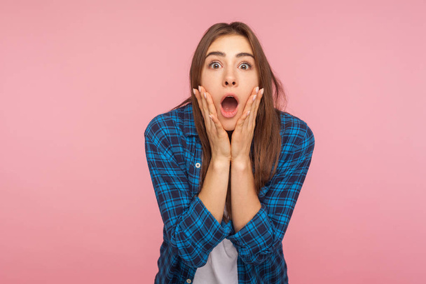 Portrait of astonished emotional funny girl in checkered shirt looking with big eyes, holding hands on face and screaming in amazement, shocked by sudden event. studio shot isolated on pink background - Photo, image
