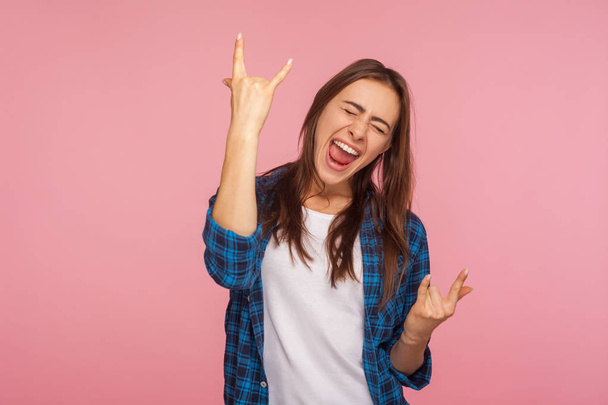 Portrait of enthusiastic overjoyed girl in checkered shirt showing rock and roll gesture and yelling with happiness, feeling to be crazy rocker on festival. studio shot isolated on pink background - 写真・画像