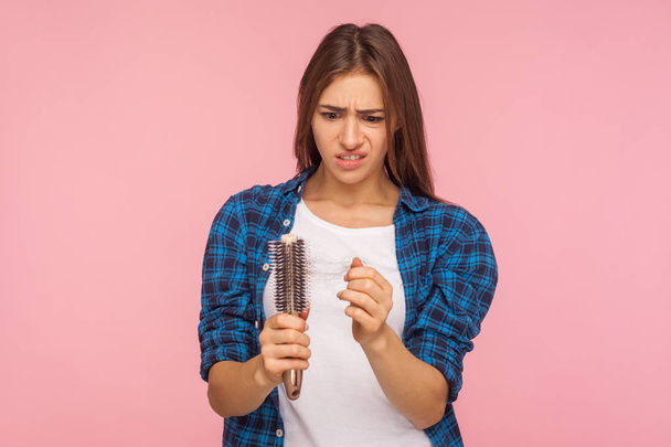 Portrait of stressed brunette woman in casual style clothes looking at unhealthy hair on brush, checking dandruff, upset by hair loss problem, alopecia. indoor studio shot isolated on pink background - Photo, Image