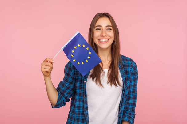 Portrait of friendly pretty woman in checkered shirt smiling broadly and holding flag of European Union, symbol of Europe, EU association and community. indoor studio shot isolated on pink background - Photo, Image