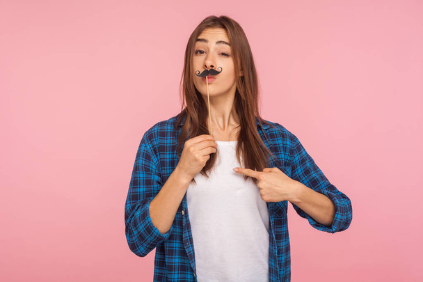 Portrait of playful girl in checkered shirt holding fake curly mustache on stick and pointing herself, having fun pretending to be bossy and arrogant. indoor studio shot isolated on pink background - Foto, Imagem