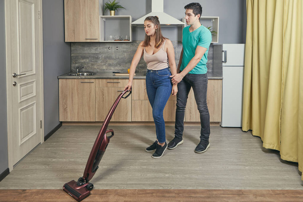 Two newlyweds had a fight. Young white man and woman in a quarrel. A 22 year old Caucasian female was offended by her male. Girl vacuums floor in kitchen, and her boyfriend distracts her from work. - Foto, Bild