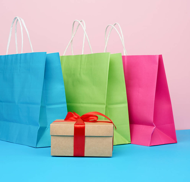 hnědá papírová krabička s dárkem a vícebarevné papírové tašky pro nakupování a dárky s bílou rukojetí stojí na růžovo-modrém pozadí - Fotografie, Obrázek