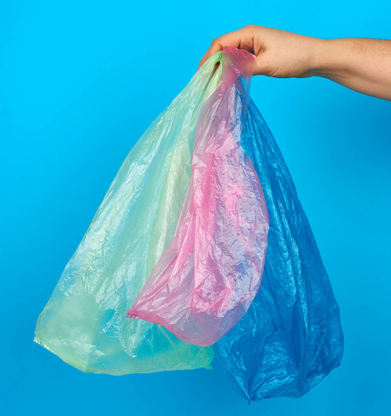 χέρι κρατά κενές πολύχρωμες πλαστικές σακούλες σε μπλε φόντο, έννοια της απόρριψης του πλαστικού και τη μετάβαση σε οικολογική συσκευασία - Φωτογραφία, εικόνα