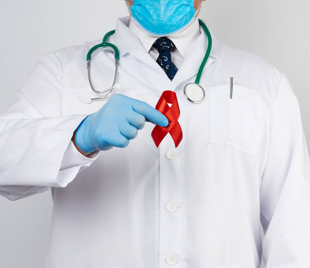 médecin en uniforme blanc et gants en latex stériles tient un ruban rouge symbole de la lutte contre la maladie SIDA et vascularite, gros plan
 - Photo, image