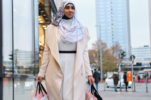 Giovane donna araba sorridente in hijab camminando felicemente per la strada della città con le borse della spesa
 - Foto, immagini