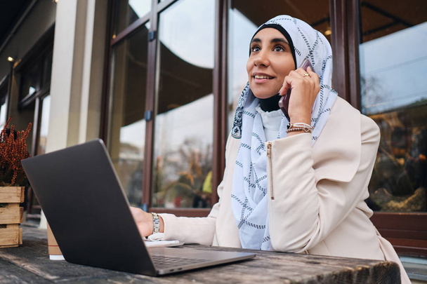 Jovem atraente mulher de negócios árabe no hijab falando no smartphone durante o trabalho no laptop na rua da cidade
 - Foto, Imagem