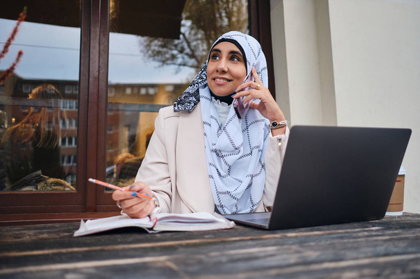 Νεαρή όμορφη Άραβα επιχειρηματίας στο χιτζάμπ ευτυχώς μιλάμε για smartphone που εργάζονται σε φορητό υπολογιστή στο δρόμο - Φωτογραφία, εικόνα