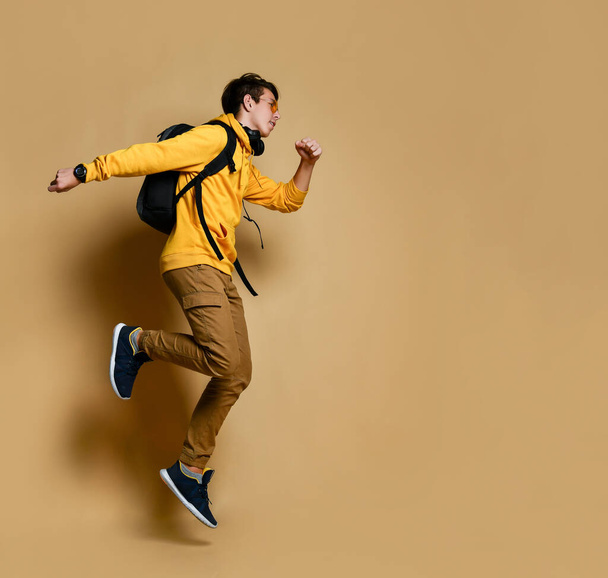 Young teen boy in comfortable clothing, sneakers and backpack jumping over yellow wall background - Фото, зображення
