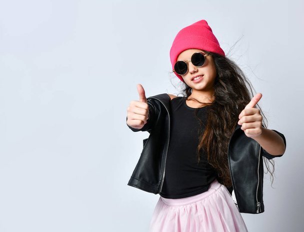 Teenage female in black leather jacket, pink hat and sunglasses. She smiling, showing thumbs up, isolated on white. Close up - Фото, изображение