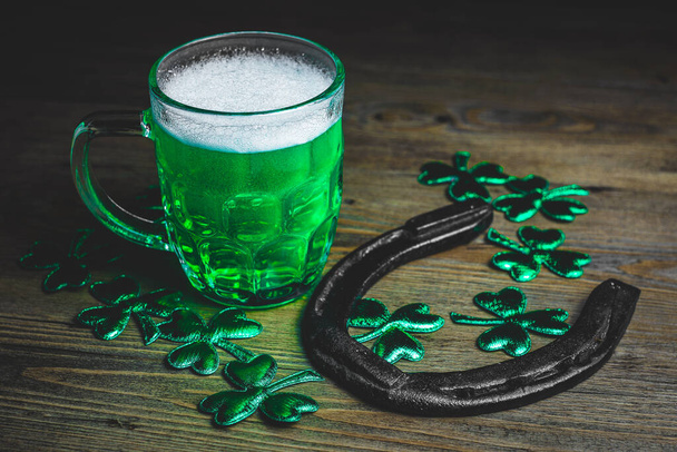 St. Patrick 's Day. Grünes Bier mit rostigem Hufeisen auf Holztisch, dekoriert mit Shamrock-Blättern. Glas Grünes Bier aus nächster Nähe. Selektiver Fokus. Unklarer Hintergrund.  - Foto, Bild