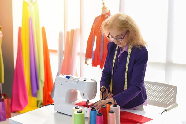 Estudio de diseño de moda que mide las tijeras de material textil que cortan la tela de la mujer mayor diseñador elegante sala de exposición colgando la máquina de coser ropa y herramientas de elaboración diseñador profesional de trabajo
 - - Foto, Imagen