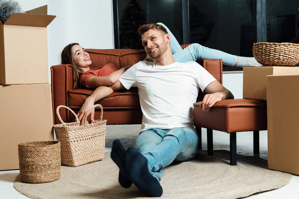 Careless young adult girlfriend lying on couch near happy boyfriend. Couple move in new apartment together and resting in living room after unpacking carton boxes - Photo, Image
