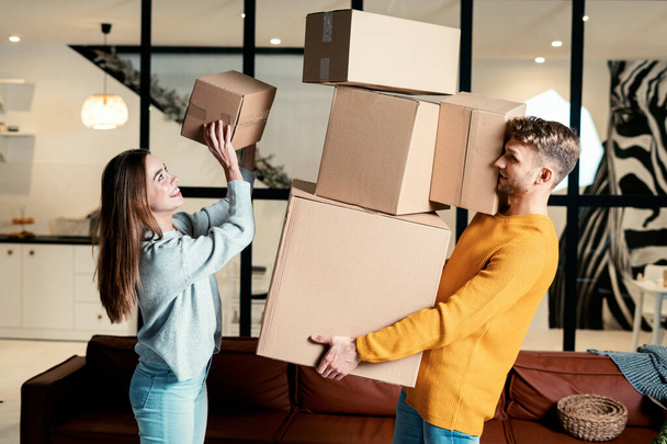 Widok z boku młodego dorosłego mężczyzny trzymającego ciężkie kartonowe pudełka w rękach, podczas gdy szczęśliwa kobieta położyła paczkę na szczycie stosu. Para razem przenieść się w nowym mieszkaniu - Zdjęcie, obraz
