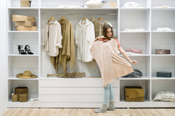 Nuori aikuinen nainen seisoo lähellä modernia vaatekaappia kotona, jossa on nykyaikainen sisustus, valitsemalla trendikkäitä vaatteita, joilla on vaikea valinta ei tiedä mitä laittaa päälle, kokeilemalla muodikasta kulumista.
 - Valokuva, kuva