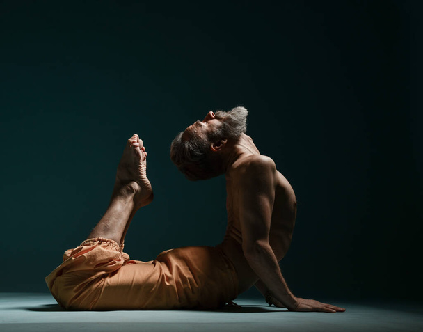 Old man with grey beard doing yoga, pilates, fitness training, stretching exercise, asana or balance workout on floor - Photo, image