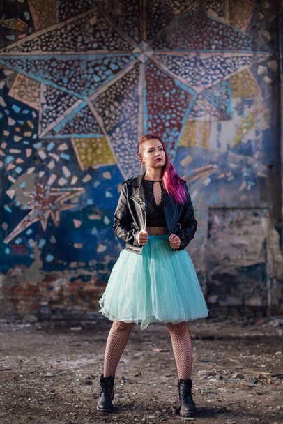Young girl with pink hair dressed in leather jacket and a blue skirt standing inside of collapsed building surrounded by ruins. Woman standing next to the old mosaic wall on the background. - Fotoğraf, Görsel