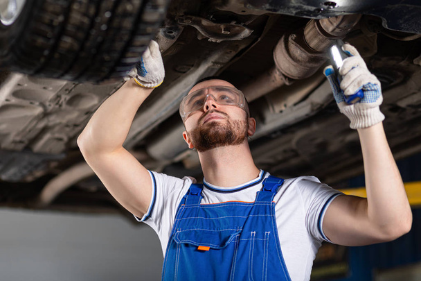 A young man auto mechanic in overalls at his workplace repairs the car's suspension, shines a lantern on the chassis. Concept Worker at an automobile repair shop, a car lifted on a lift - Photo, Image