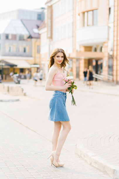 Cute pretty girl with professional makeup in a pink top and denim skirt in a summer city holding a bouquet of white and pink roses. - Photo, Image