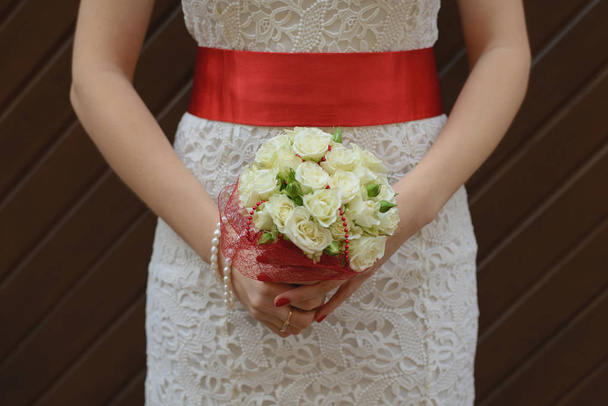 Λευκό γαμήλιο μπουκέτο με τριαντάφυλλα στα χέρια της νύφης, ένα κορίτσι με λευκό φόρεμα με κόκκινη σατέν κορδέλα στα χέρια της. - Φωτογραφία, εικόνα