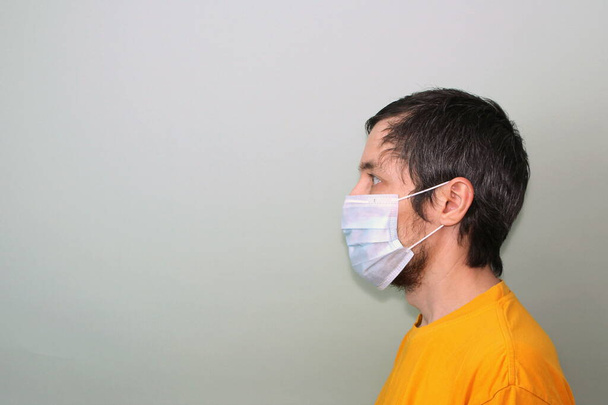 Middle-aged adult man in a yellow T-shirt on a gray background in a medical mask sideways in profile. Disease protection coronavirus epidemic problem from China. - Foto, imagen