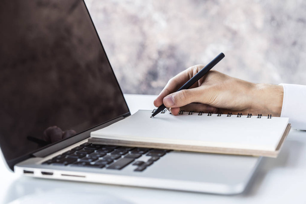 Üzletember ír egy tollal a naplójában a laptop billentyűzetén egy napfényes irodai, üzleti és oktatási koncepcióban. Közelről. - Fotó, kép