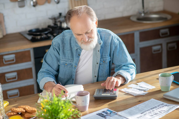 Bearded grey-haired man sitting at the table and making calculations - Foto, Imagem