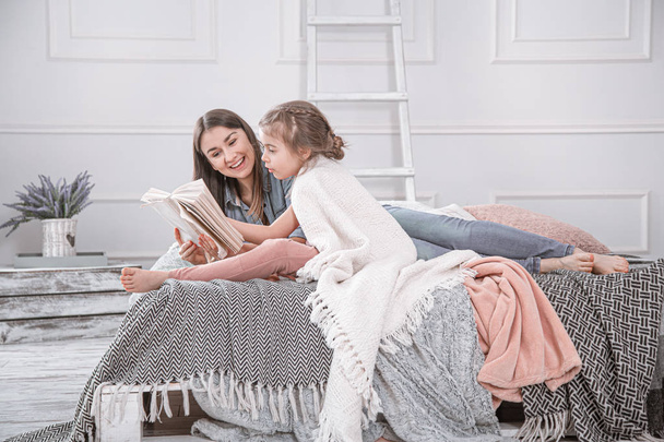 Portrait of a smiling young cute mother and daughter reading a book lying and relax in the bed in a bright big white room. Concept of children's education and development. - Fotó, kép