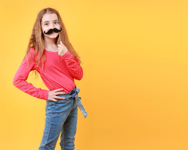 Νεαρό κορίτσι με μαύρο χάρτινο μουστάκι που διασκεδάζει, παιδικό πορτραίτο σε κίτρινο φόντο, αστεία έννοια.Κενό αντίγραφο χώρου.Καλή διασκέδαση τρόπο ζωής. - Φωτογραφία, εικόνα