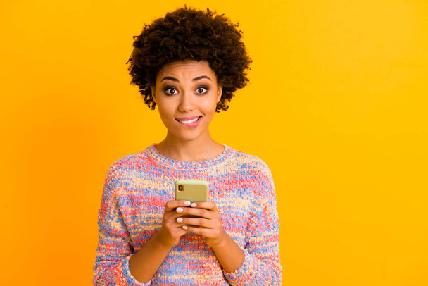 Portrait of positive dreamy inspired afro american girl use cellphone bite lips wait likes posts subscribers followers repost comments wear spring jumper isolated yellow color background - Zdjęcie, obraz