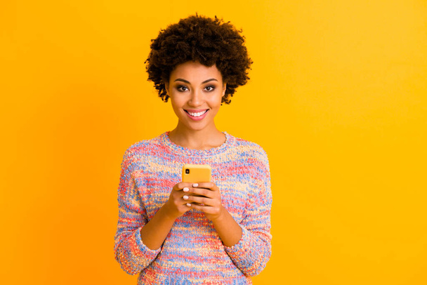 Portrait of positive cheerful afro american girl use smart phone enjoy following post reposting news share social media information wear stylish outfit isolated over yellow color background - Fotografie, Obrázek