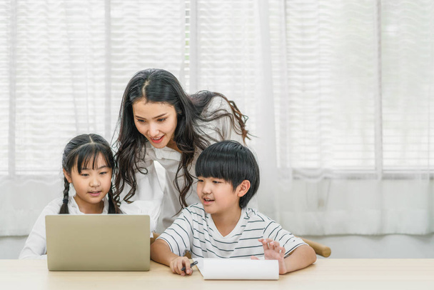 Young Asian Single Mother teaching or helping daughter and son doing home work with technology laptop in home, home school education, relationship and family, communication and conversation concept - Foto, immagini
