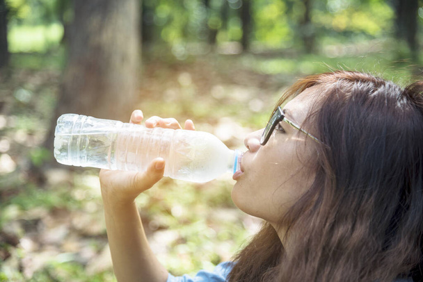 Zdrowa kobieta trzymająca wodę mineralną w dłoniach na zewnątrz miasta w zielonym parku uśmiecha się i patrzy w kamerę ze szczęśliwą twarzą. Młoda kobieta szczęśliwy outdoor styl życia trzymając butelkę wody. - Zdjęcie, obraz