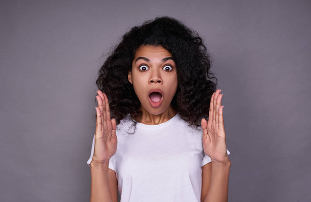 Curly optimistic woman raises palms from joy, happy to receive awesome present from someone, shouts loudly, dressed in casual white t shirt, isolated on grey background. Excited Afro female yells. - Photo, Image