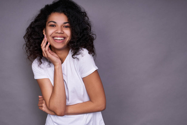 A cheerful African American with a pleasant smile, curly dark hair, healthy skin, wears a white T-shirt, stands on a gray background, in a good mood, smiles a snow-white smile and looks straight. - Foto, afbeelding