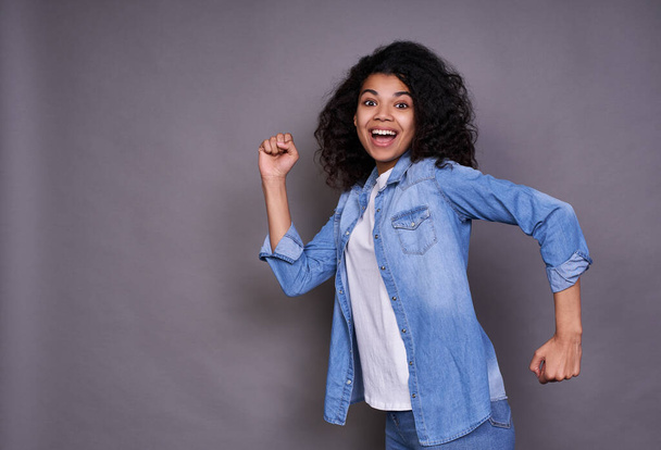 Portrait of a cheerful young African-American girl, a joyful thousand-year-old African-American girl walks through the air, runs to the left on a gray studio background, smiles at the camera. - Foto, Bild