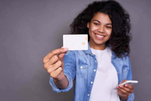 Retrato de uma jovem africana excitada, com um penteado encaracolado fofo, em uma camiseta branca e camisa jeans, mostrando um cartão de crédito de plástico enquanto segura um telefone celular isolado em um fundo cinza
. - Foto, Imagem