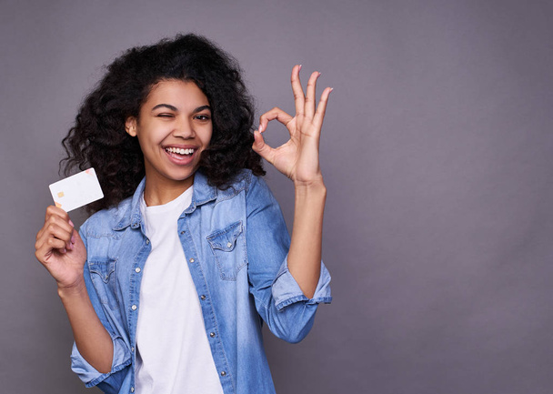 Portrait of a smiling young beautiful curly african american woman, in a denim shirt holding credit cards, shows her okay sign with her left hand. Online shopping concept, user lending. - Foto, afbeelding