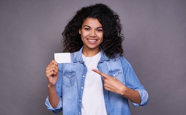 Portrait of a smiling young beautiful curly african american woman holding and pointing fingers at credit cards isolated on gray background. Online shopping concept, user lending. - Фото, изображение