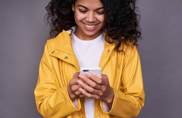 Cropped frame, of a happy young African girl, student, with a lush wavy hairstyle, in a yellow raincoat using a mobile phone, writes reads SMS of her beloved. Modern technology concept. - Foto, Imagem