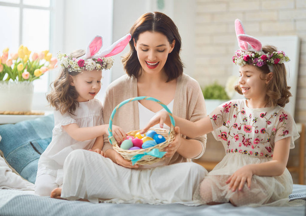 Happy holiday! Mother and her daughters with painting eggs. Family celebrating Easter. Cute little children girls are wearing bunny ears.                                                               - Foto, Imagem