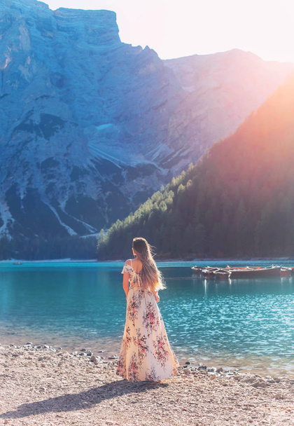 Girl with hair flying in wind red-haired, long silk elegancy dress floral print turned away stands on shore Lake Bryce enjoy silence beauty sunshine mountains. Backdrop boat water mountain with forest - Photo, Image