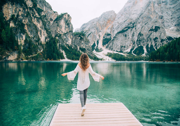 Romantic girl with long hair loose, turned away, walk spread hands on wooden pier Lake Braies. Tourist enjoy mysterious nature amazing Alpine mountains with green forest clear water silence relaxation - Photo, Image
