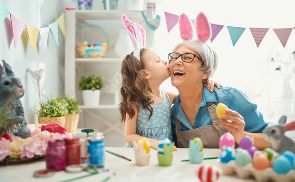  Happy easter! A grandmother and her granddaughter painting Easter eggs. Happy family preparing for Easter. Cute little child girl wearing bunny ears on Easter day. - Фото, изображение