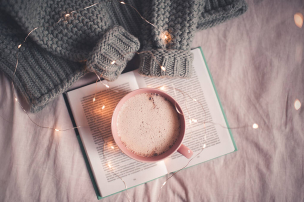 ベッドでコーヒーブレイク。ニットセーターを着たままオープンブックに滞在するラテのカップを閉じます。おはようございます。冬の季節.  - 写真・画像
