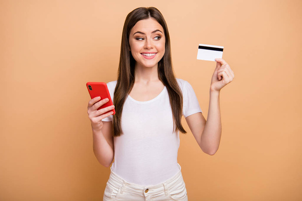Portrait of positive cheerful girl hold look debit card want online shopping on cellphone connection check her income earnings online wear pants trousers isolated beige color background - Valokuva, kuva